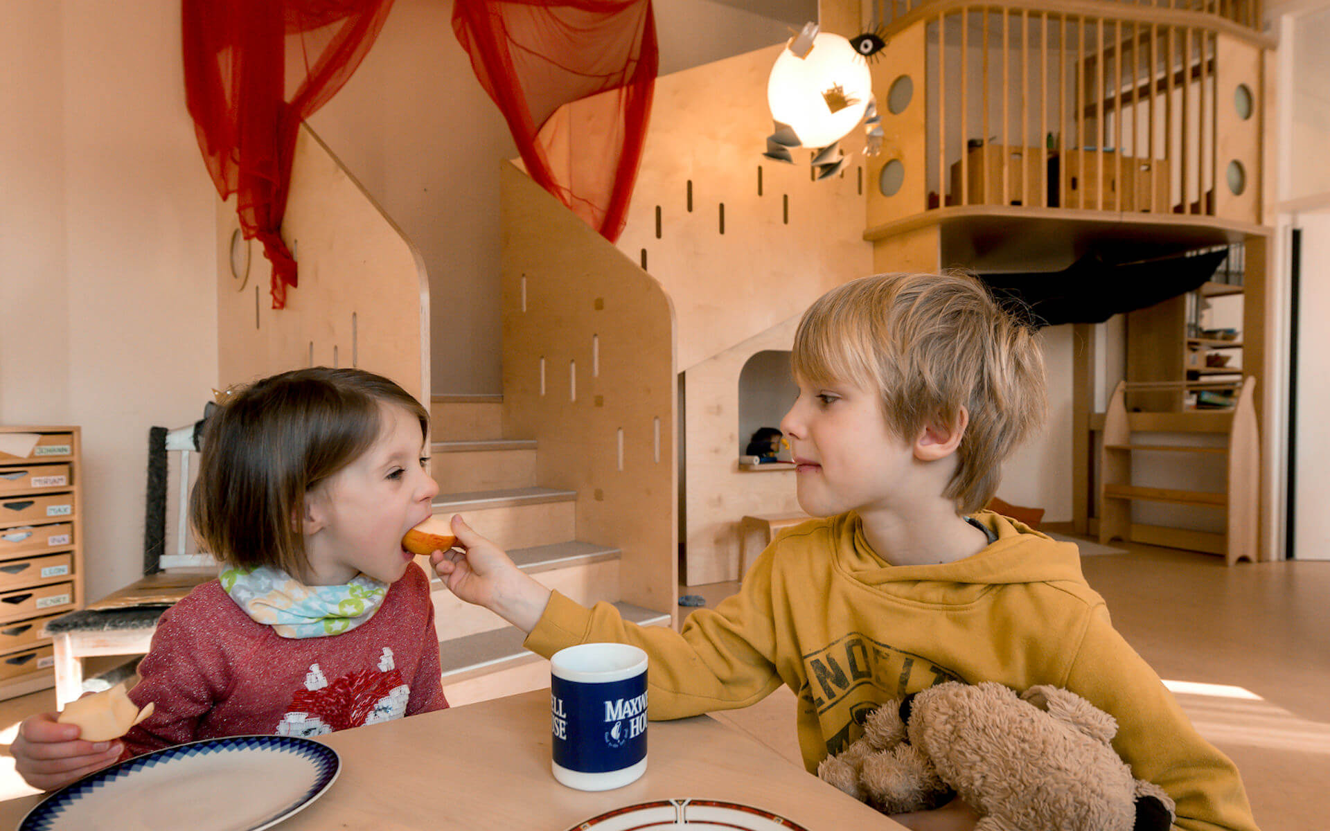 Zwei Kinder beim gemeinsamen Essen im Kinderrestaurant, im Hintergrund ein Podest aus Holz